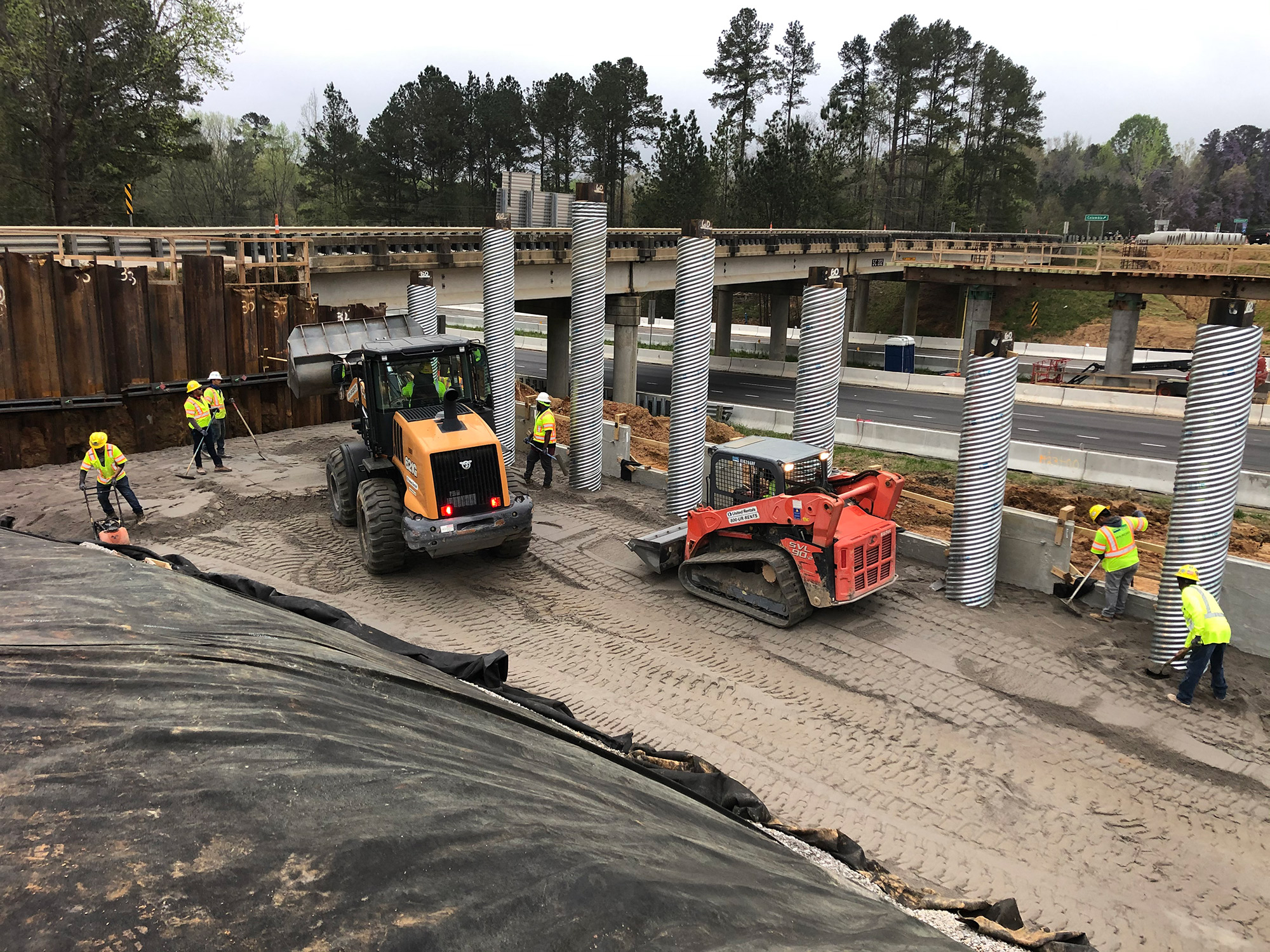 Men and equipment placing backfill for the retaining wall at the SC-202/Exit 85 overpass.  Steel piles that will support the future bridge are protected with spiral ribbed steel cans during the placement of the select backfill material.